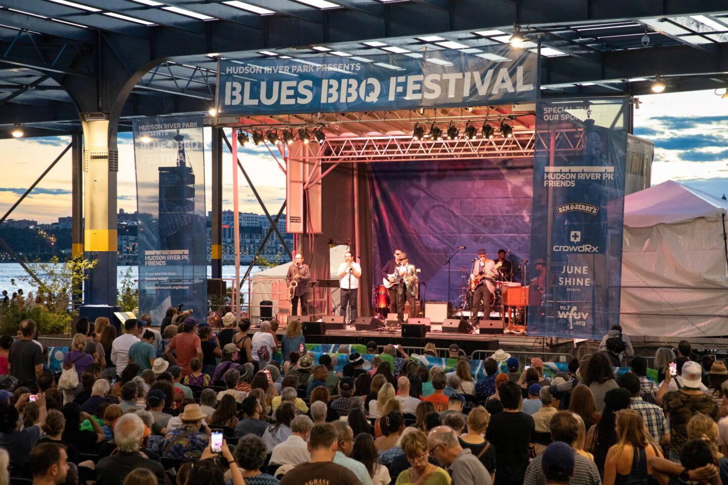 A band performs on the Hudson River Park Blues BBQ Festival stage at sunset while a crowd watches on