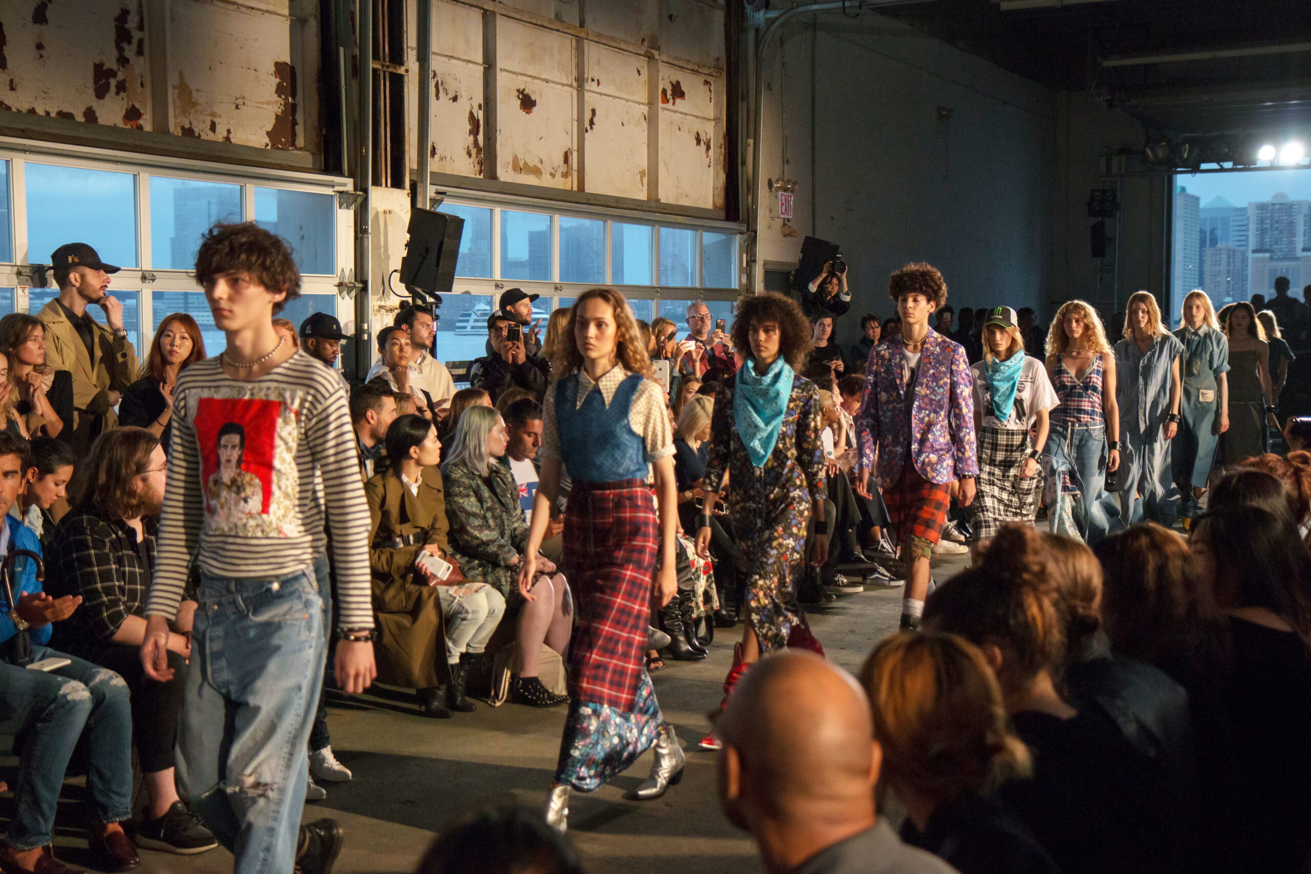 Models walk down a fashion show runway with audience members on either side